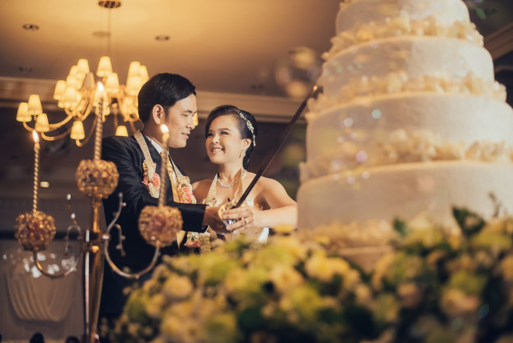 คู่บ่าวสาวชาวไทยกำลังเฉลิมฉลองในระหว่างงานแต่งงานที่มีการตกแต่งอย่างหรูหราพร้อมชั้นเค้กขนาดใหญ่