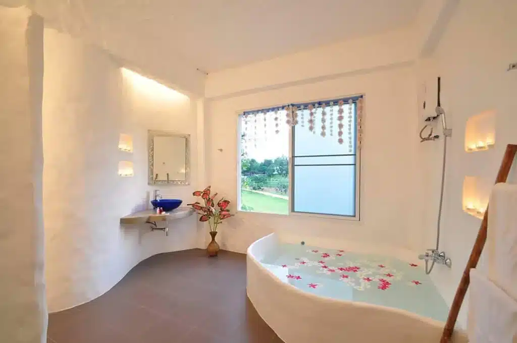 ห้องน้ำสว่างสดใสพร้อมอ่างอาบน้ำเข้ามุมประดับด้วยกลีบดอกไม้ อ่างล้างมือสีฟ้า และวิวเขียวขจีผ่านหน้าต่าง รีสอร์ทมวกเหล็็ ที่พักมวกเหล็กติดน้ำตก
