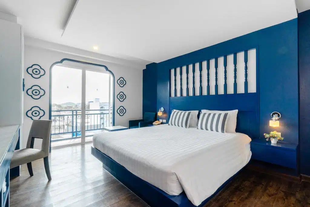 ห้องนอนร่วมสมัยที่มีเตียงขนาดใหญ่พร้อมผ้าปูที่นอนสีขาวตัดกับผนังสีฟ้า ตกแต่งโดยได้รับแรงบันดาลใจจากถนนคนเดินภูเก็ต และ ถนนคนเดินภูเก็ต