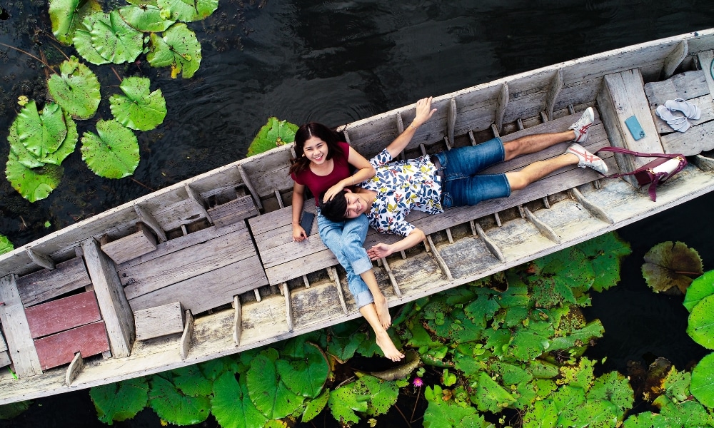 คนสองคนพักผ่อนบนเรือไม้ที่รายล้อมไปด้วยดอกบัวท่ามกลางความงามตามธรรมชาติของนครปฐม นครปฐมที่เที่ยวธรรมชาติ