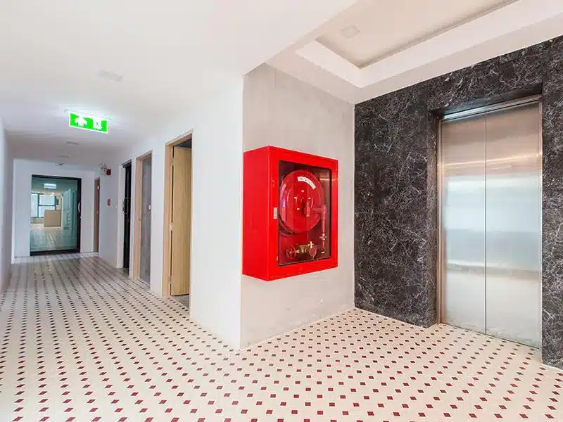โถงทางเดินในอาคารสมัยใหม่มีตู้ม้วนสายยางดับเพลิงสีแดงและประตูลิฟต์ที่ตัวต้านทานริมโขงดามุกหาร ที่พักริมโขงมุกดาหาร