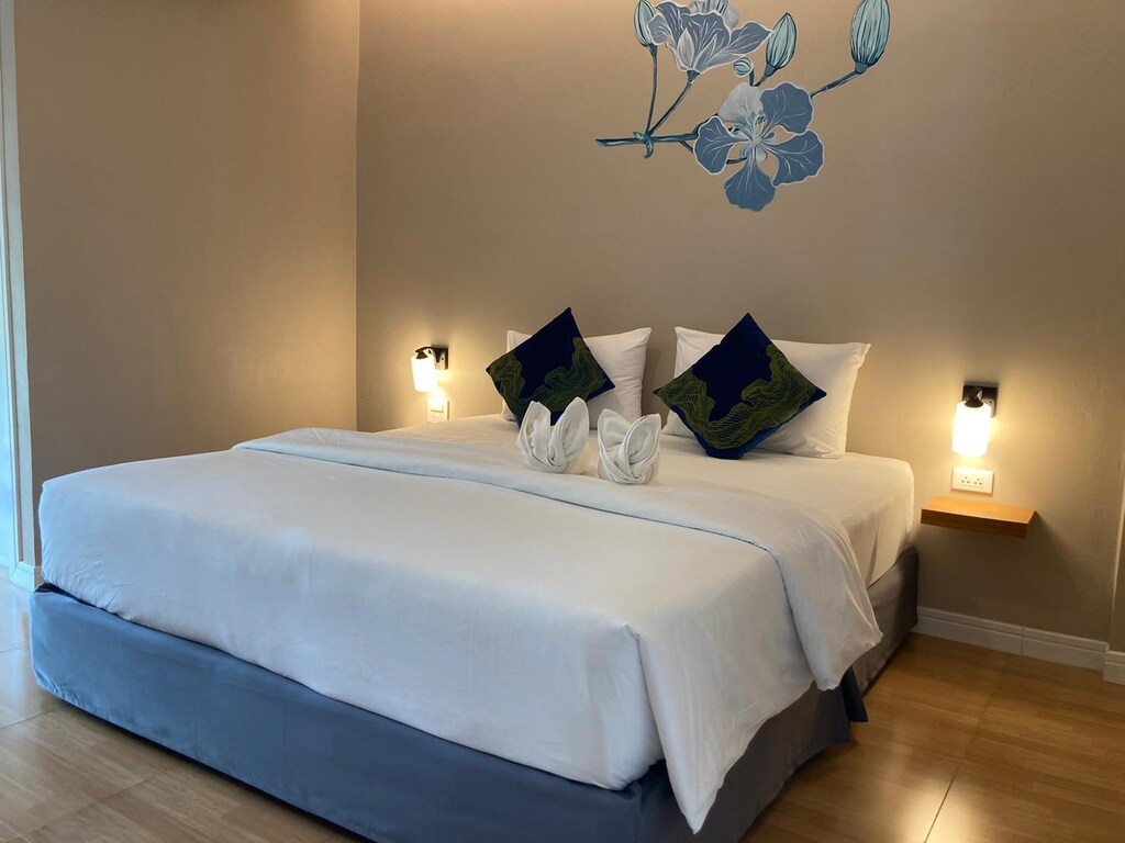 เตียงที่ตกแต่งอย่างประณีตด้วยผ้าปูที่นอนสีขาว เน้นสีฟ้า และหมอนตกแต่งในห้องที่มีแสงไฟนวลตาและดีไซน์ผนังลายดอกไม้ สถานที่ท่องเที่ยวสกลนคร