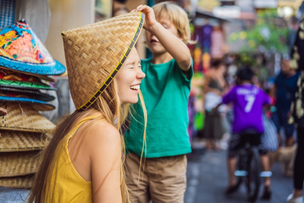 ผู้หญิงสวมหมวกทอใบใหญ่ยิ้มแย้ม ขณะที่เด็กๆ ยืนอยู่ข้างหลัง ถือหมวกให้เข้าที่ระหว่างเยือนราชบุรี ที่เที่ยวบาหลี