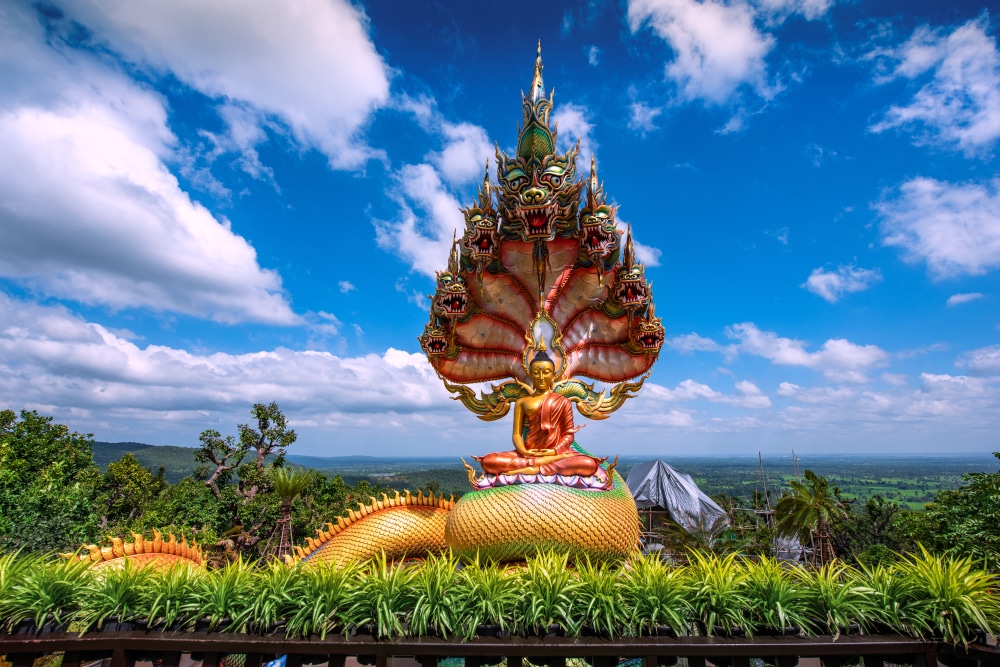 รูปปั้นพระพุทธรูปสีสันสดใสที่มีหลังคางูหลายเศียร ตั้งตระหง่านอยู่บนท้องฟ้าสีครามและมีเมฆปุย ที่เที่ยวสกลนคร