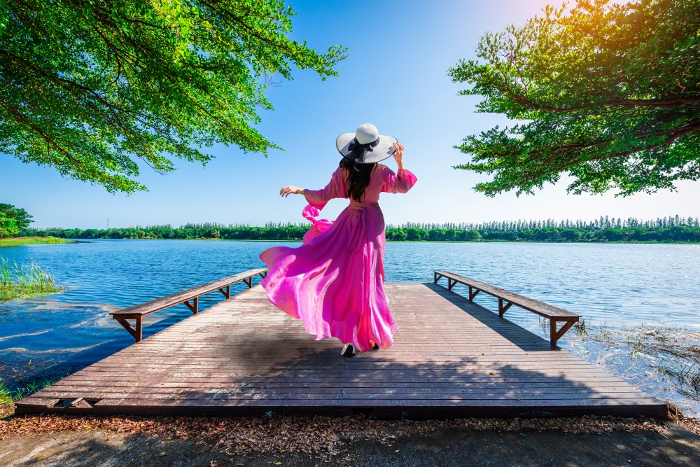 ผู้หญิงในชุดสีชมพูและหมวกกันแดดกำลังเพลิดเพลินกับวันที่มีแสงแดดสดใสบนท่าเทียบเรือไม้ริมทะเลสาบ ที่เที่ยวสกลนคร