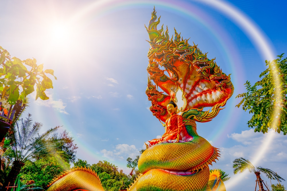 รูปปั้นพระพุทธรูปทองคำประทับนั่งบนงูหลากสีสันภายใต้แสงอาทิตย์ที่สดใสและมีรัศมีสีรุ้ง ที่เที่ยวสกลนคร