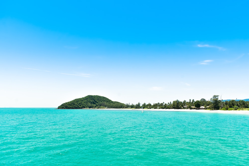 ชายหาดเขตร้อนที่มีท้องฟ้าสีฟ้าใสและน้ำทะเลสีฟ้าคราม มีเกาะเล็กๆ อยู่ไกลๆ ที่เที่ยวสมุย