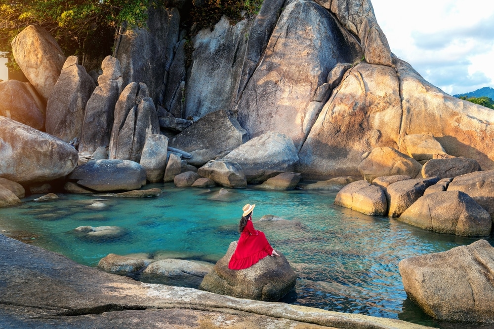 ผู้หญิงในชุดสีแดงนั่งอยู่บนโขดหินริมสระน้ำบนภูเขาอันเงียบสงบ ที่เที่ยวเกาะสมุย ที่เที่ยวเดือนเมษายน