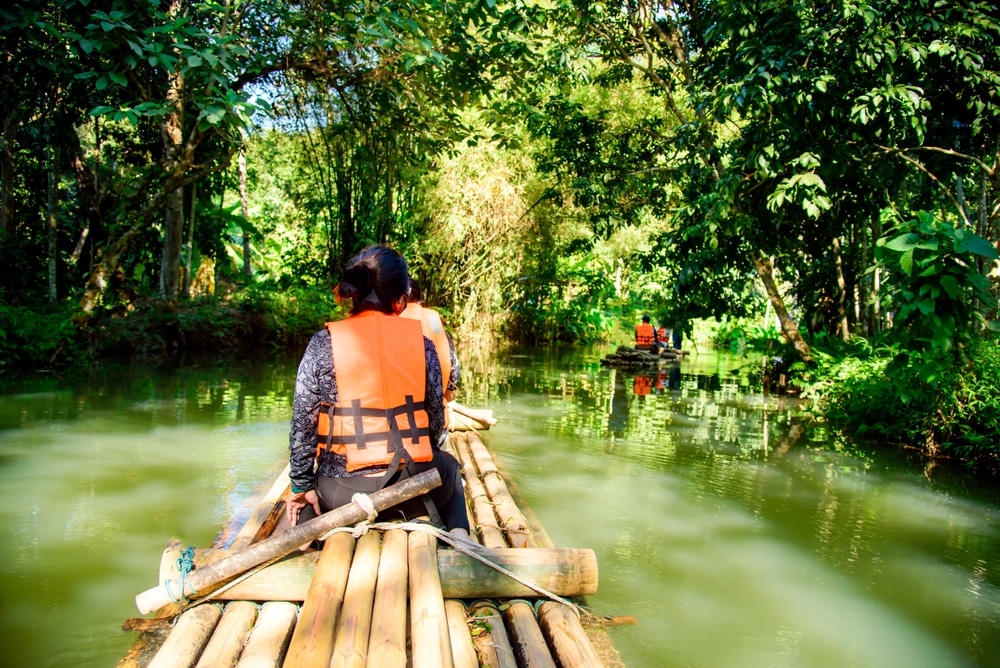 คนสวมเสื้อชูชีพกำลังพายเรือไม้ไผ่ในแม่น้ำอันเงียบสงบท่ามกลางแมกไม้เขียวขจีในการท่องเที่ยวเดือนเมษายน ที่เที่ยวเดือนเมษายน