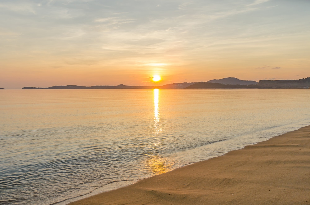 พระอาทิตย์ตกเหนือผืนน้ำอันเงียบสงบพร้อมแสงสะท้อนสีทองใกล้หาดทราย ที่เที่ยวเกาะสมุย