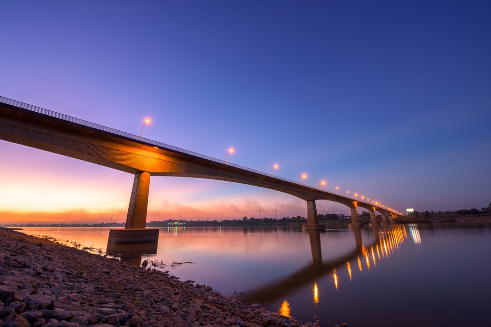 สะพานสว่างไสวเหนือผืนน้ำอันเงียบสงบในเวลาพลบค่ำ บั้งไฟพญานาค ประดับประดาโดยบั้งไฟพญานาค