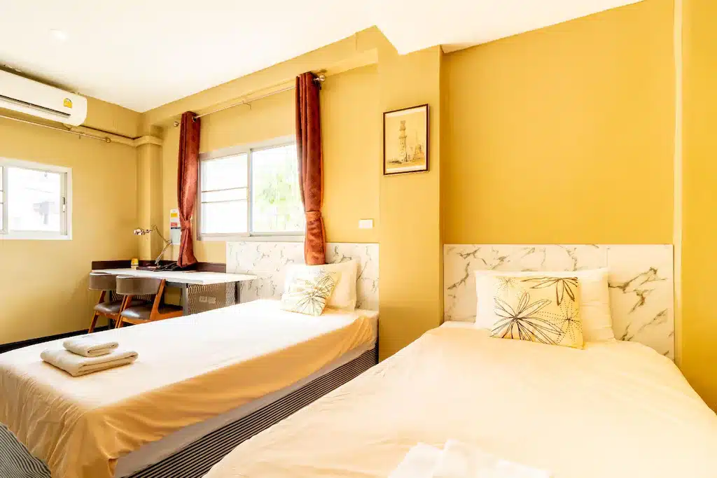 ห้องนอนแฝดสว่างและสะอาดพร้อมผนังสีเหลือง ผ้าปูเตียงสีขาว และโต๊ะเล็กริมหน้าต่าง ใช้เวลาใกล้กทม. ที่พักใกล้กรุงเทพ