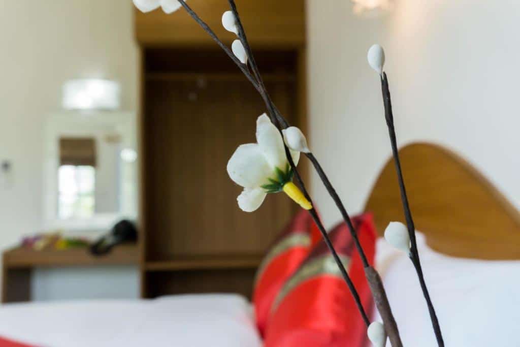 ภาพระยะใกล้ของดอกไม้สีขาวบนกิ่งก้าน โดยมีพื้นหลังเบลอซึ่งมีเตียงที่จัดอย่างประณีตและหมอนสีแดงในห้องที่สว่างสดใสของรบกวน ที่พักจังหวัดตาก ตาก