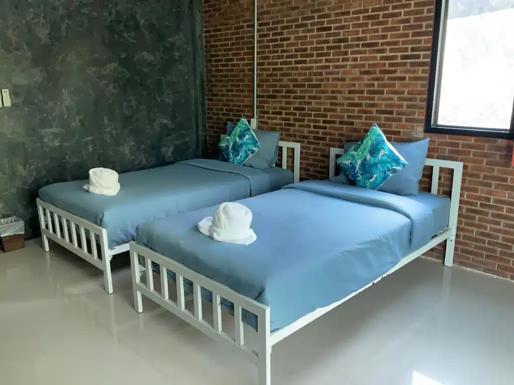 เตียงเดี่ยว 2 เตียงพร้อมผ้าปูที่นอนสีฟ้าในห้องที่ชัยนาทรีสอร์ท มีผนังอิฐและปูนสีเข้มและมีหน้าต่างบานใหญ่ ที่พักชัยนาท