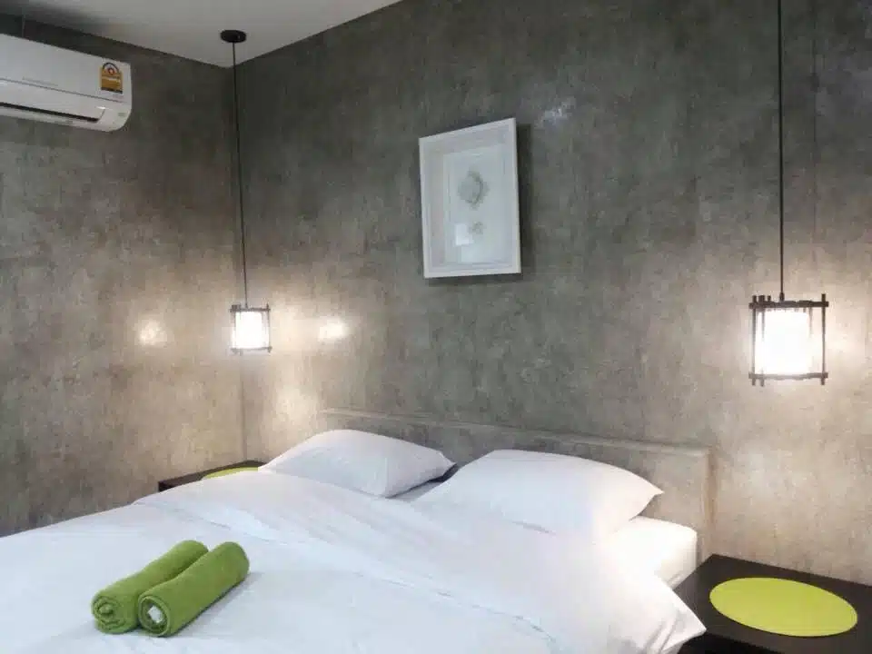 ห้องนอนทันสมัยในโรงแรมตาก มีเตียงคู่พร้อมชุดเครื่องนอนสีขาว ผ้าเช็ดตัวสีเขียว 2 ผืน โคมไฟแขวนเพดาน และผนังสีเทาที่มีพื้นผิว โรงแรมจังหวัดตาก