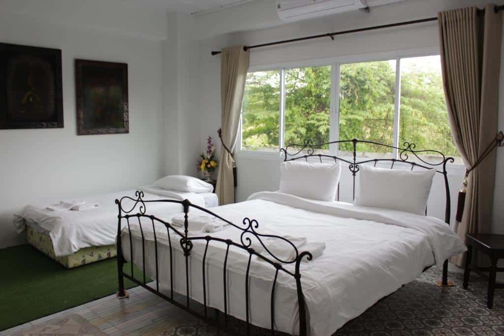 ห้องพักในโรงแรมที่สว่างและโปร่งสบายพร้อมเตียง 2 เตียงที่มีโครงเหล็กดัด ชุดเครื่องนอนสีขาว หน้าต่างบานใหญ่พร้อมผ้าม่าน และภาพวาดเหนือโต๊ะข้างเตียงในตอนกลางคืน ที่พักจังหวัดตาก