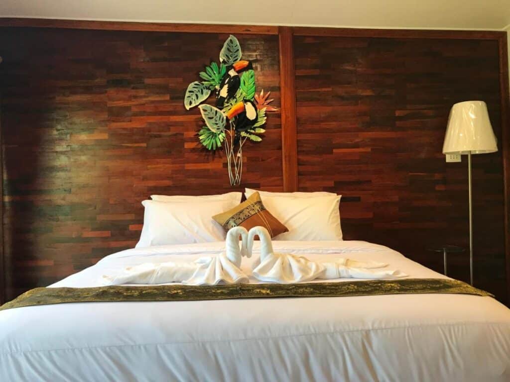 เตียงที่ตัดเย็บอย่างปราณีตด้วยผ้าปูเตียงสีขาวในห้องที่มีผนังไม้และผนังสีสันสดใสเหนือเตียงในราชบุรี ที่เที่ยวบางกระเจ้า
