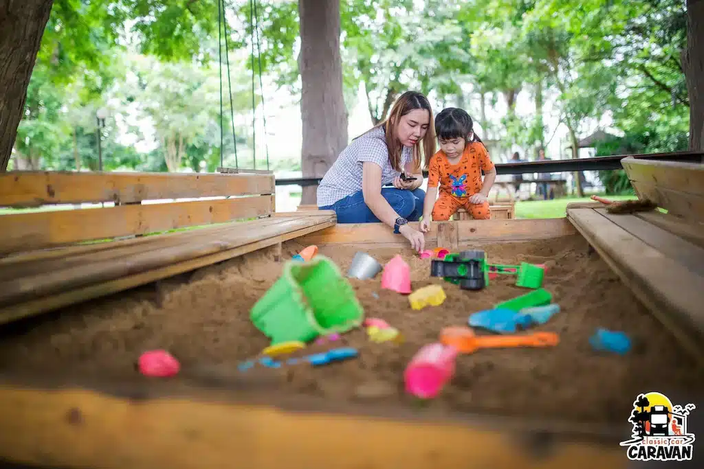 ผู้หญิงและเด็กเล็กเล่นด้วยกันในกล่องทรายที่เต็มไปด้วยของเล่นสีสันสดใสที่สวนสาธารณะใกล้รีสอร์ทชัยนาท โรงแรมชัยนาท