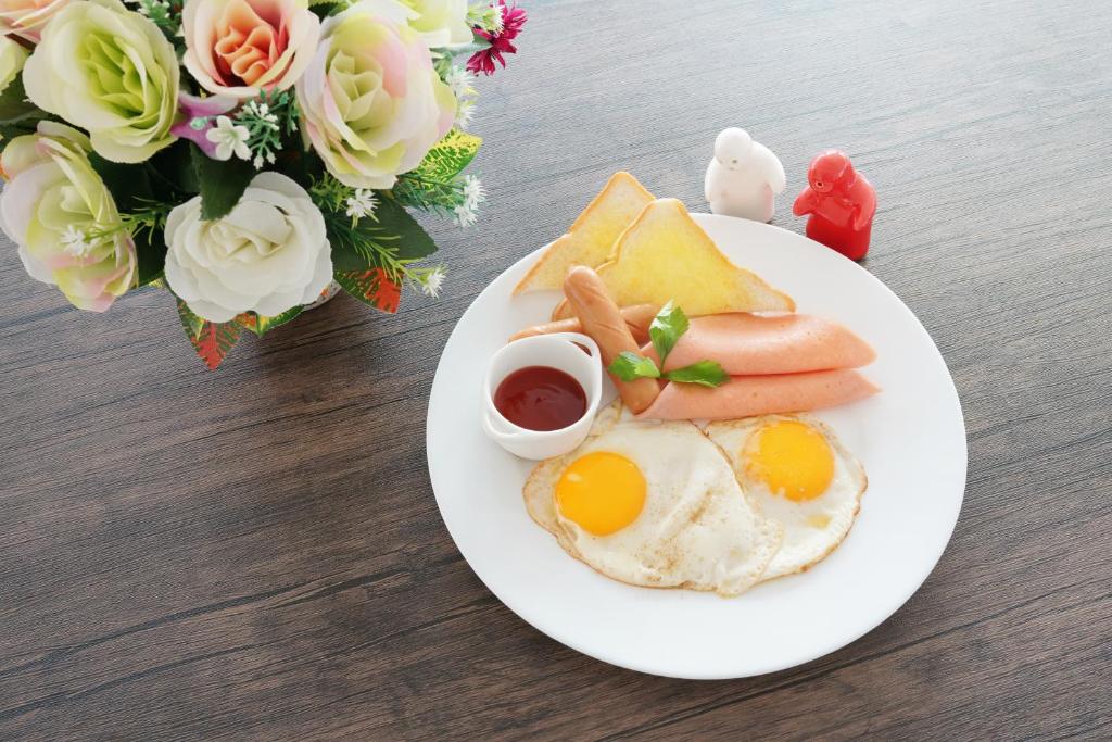 จานอาหารเช้าที่มีไข่แดดเดียว ขนมปังปิ้ง ไส้กรอก และซอสมะเขือเทศ ข้างๆ ช่อดอกไม้และของตกแต่งบนโต๊ะไม้ที่ร้านนี้มีสกลนค ที่พักสกลนคร