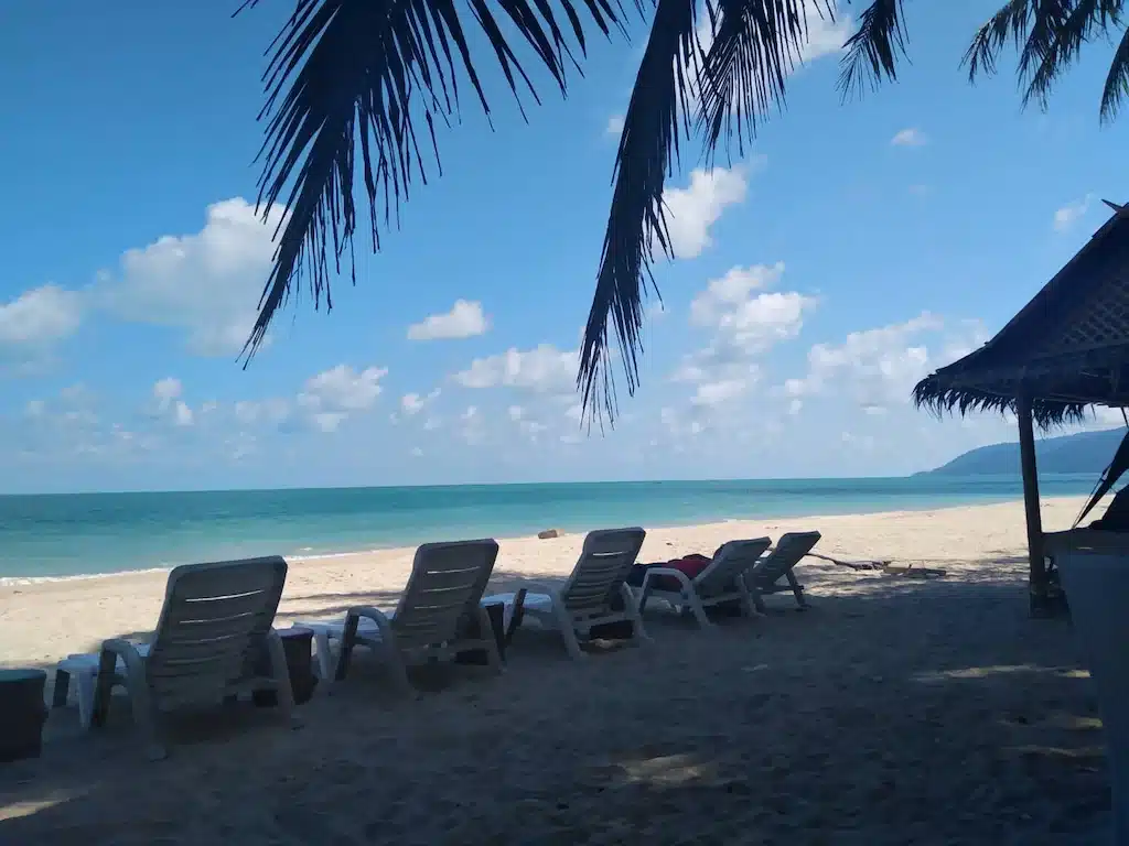 ฉากชายหาดที่มีเก้าอี้นั่งเล่นว่างๆ ใต้ต้นปาล์ม มองเห็นวิวทะเลสีฟ้าและท้องฟ้าใสๆ ในน่านที่เที่ยว ขนอมที่พัก