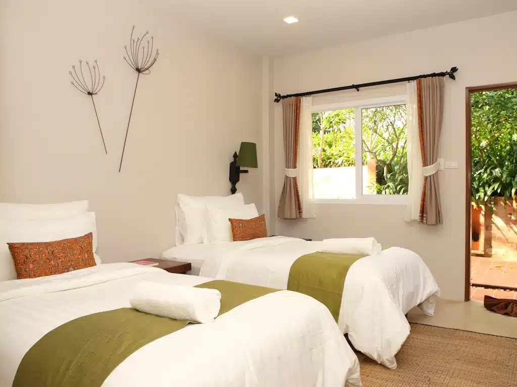 ห้องนอนสว่างสดใสและเป็นระเบียบเรียบร้อยพร้อมเตียงแฝด 2 เตียง ผ้าห่มสีเขียว ผ้าปูที่นอนสีขาว ที่พักเกาะพยาม และผ้าม่านตกแต่งบนหน้าต่างที่มองเห็นความเขียวขจีที่ Blue Sky Resort Ko Phayam