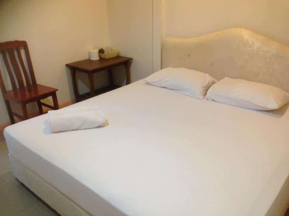 ห้องนอนเรียบง่ายที่ชัยนาทรีสอร์ทมีเตียงขนาดใหญ่พร้อมผ้าปูที่นอนสีขาว เก้าอี้ไม้ และโต๊ะข้างเตียงขนาดเล็กพร้อมต้นไม้ โรงแรมชัยนาท