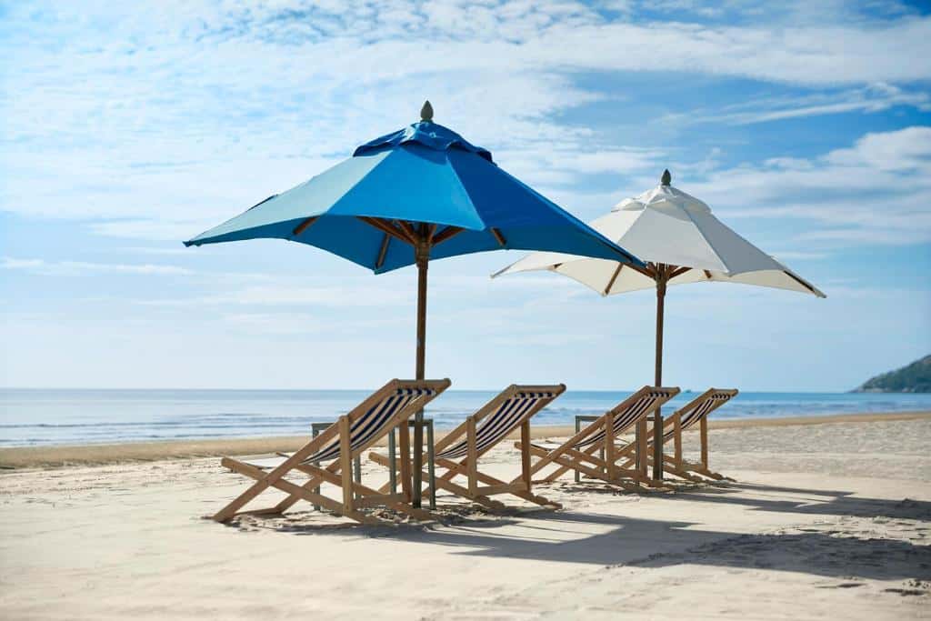 เก้าอี้ชายหาดไม้สี่ตัวใต้ร่มสองคันบนหาดทรายในหัวหิน โดยมีท้องฟ้าแจ่มใสและทะเลอันเงียบสงบเป็นฉากหลัง ที่พักหัวหิน