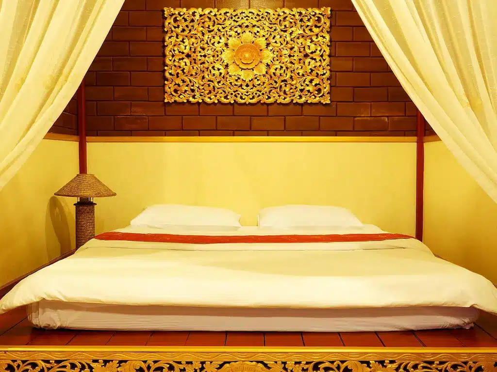 ห้องนอนแบบดั้งเดิมที่มีเตียงไม้สี่เสาพร้อมผ้าปูที่นอนสีขาวและผนังตกแต่งด้วยลายดอกไม้สีทองด้านบน ขนาบข้างด้วยผ้าม่านสีขาวในบรรยากาศเงียบสงบ โรงแรมจังหวัดตาก