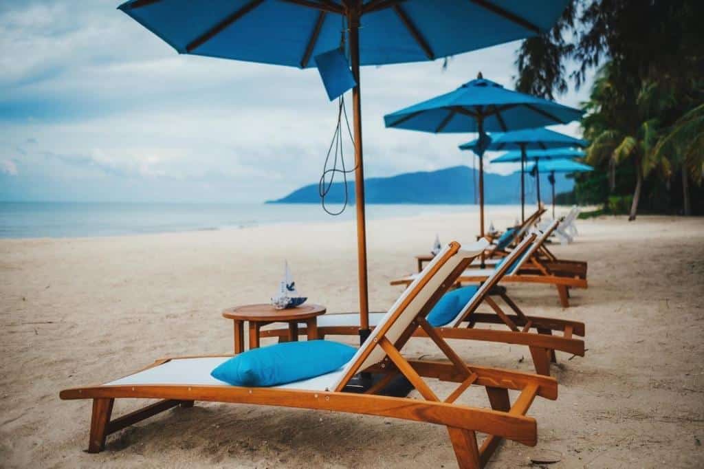 เก้าอี้ไม้นั่งเล่นเป็นแถวและร่มสีฟ้าบนหาดทรายในจังหวัดน่าน หันหน้าไปทางมหาสมุทรภายใต้ท้องฟ้าที่มีเมฆมาก อ