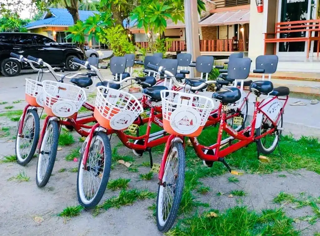 จักรยานเรียงกันเป็นแถวสีแดงพร้อมตะกร้าสีขาวจอดอยู่บนทางเดิน โดยมีต้นไม้และที่พักริมทะเลเป็นฉากหลัง ที่พักขนอมแบบครอบครัวติดทะเล