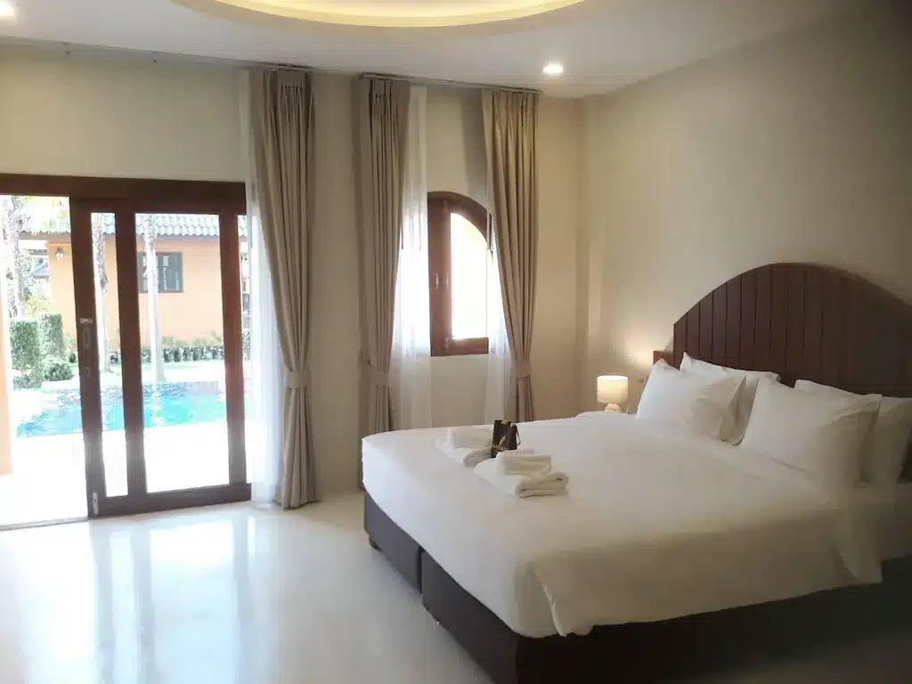 ห้องพักโรงแรมในน่านมีเตียงขนาดใหญ่พร้อมผ้าปูเตียงสีขาว หัวเตียงไม้ และประตูบานเลื่อนที่ทอดไปสู่สระน้ำ โรงแรมขนอม
