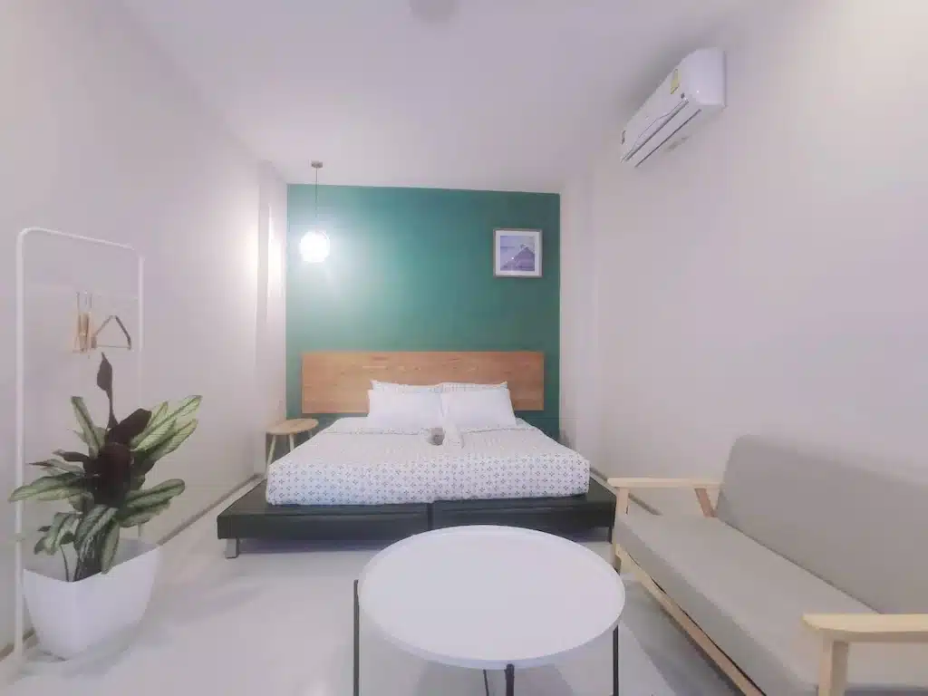ห้องนอนเล็กทันสมัยพร้อมเตียงไม้ ชุดเครื่องนอนสีขาว โซฟา โต๊ะกลม ผนังสีเขียว และเครื่องปรับอากาศที่มองเห็นวิวริมแม่น้ำฉะเชิงเทรา ที่พักฉะเชิงเทราริมน้ำ