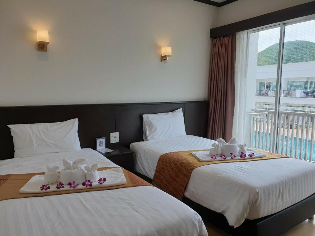 ที่พักหาดเตยงาม ห้องพักในโรงแรมทันสมัยที่มีเตียงเดี่ยว 2 เตียงพร้อมผ้าปูที่นอนสีขาวและดอกไม้สีม่วงด้านบน โคมไฟติดผนัง และหน้าต่างที่มองเห็นวิวระเบียงพร้อมวิวสระว่ายน้ำที่หาดหาด