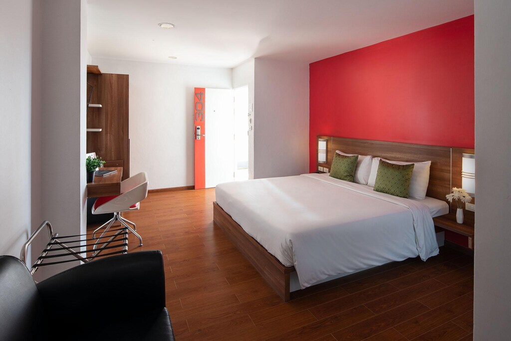 ห้องนอนทันสมัยพร้อมเตียงขนาดใหญ่ตัดกับผนังเน้นสีแดง พื้นไม้ และพื้นที่นั่งเล่นเล็กๆ พร้อมเก้าอี้สี ที่พักจังหวัดตาก ขาวและโต๊ะเหล็กสีดำในโรงแรมตา