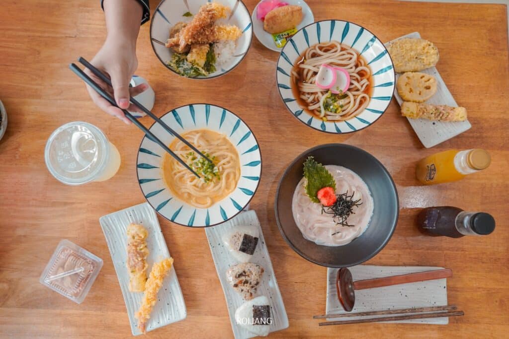 มุมมองด้านบนของโต๊ะทานอาหารร้านอาหารญี่ปุ่นในภูเก็ตที่มีอาหารหลากหลายทั้งบะหมี่ ข้าวหน้าเทมปุระ และเครื่องดื่ม มองเห็นมือของบุคคลหยิบอาหารด้วย  ร้านอาหารญี่ปุ่นภูเก็ต