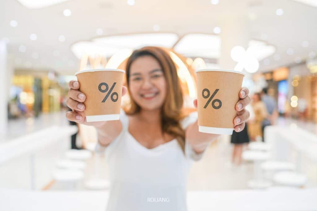 ผู้หญิงยิ้มถือถ้วยกาแฟสองใบที่มีโลโก้สัญลักษณ์เปอร์เซ็นต์ ยืนอยู่ในร้านอาหารเซ็นทรัลภูเก็ตที่มีแสงสว่างจ้า ร้านอาหารในเซ็นทรัลภูเก็ต