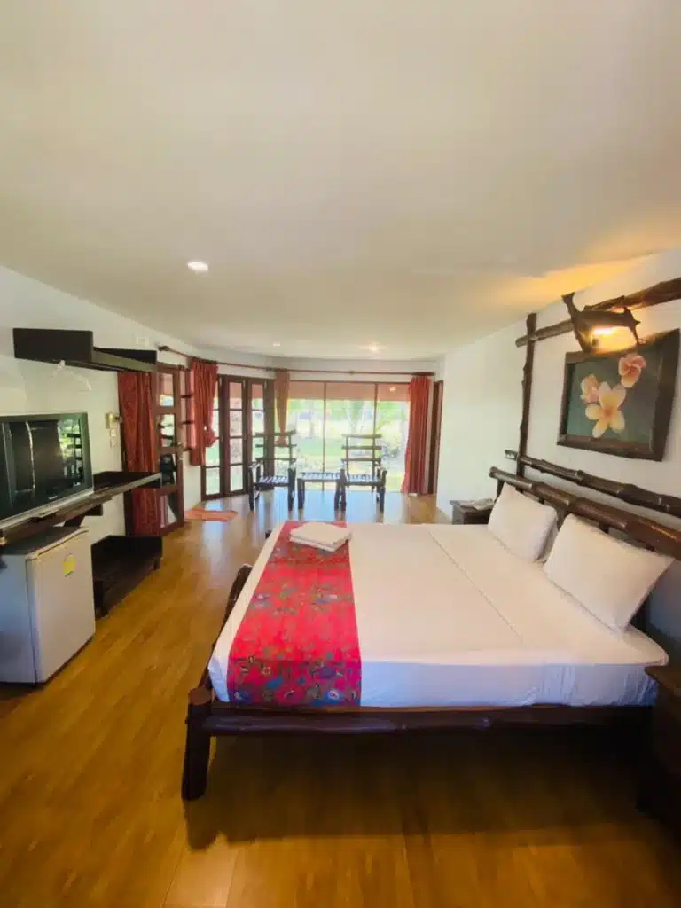 ห้องพักในโรงแรมแสนสบายที่มีเตียงขนาดใหญ่พร้อมผ้าปูเตียงสีขาวและผ้าคลุมสีสันสดใส พื้นไม้ และพื้นที่รับประทานอาหารในพื้นหลังพร้อมแสงธรรมชาติกรองผ่านม่านโปร่งในน่านที่เ โรงแรมขนอม