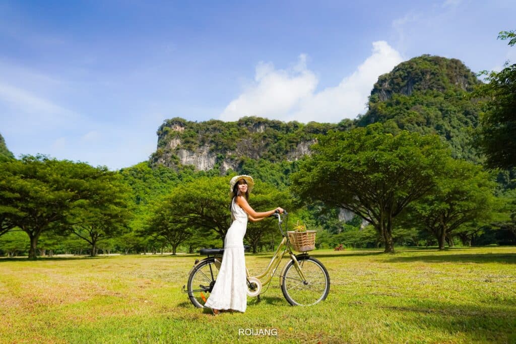 หญิงสาวในชุดสีขาวและหมวกโพสท่ากับจักรยานในทุ่งหญ้าที่รายล้อมไปด้วย พูลวิลล่าพังงา ต้นไม้สีเขียวชอุ่มและภูเขา ชวนให้นึกถึงความงามอันเงียบสงบใกล้กับ พูลวิลล่าพังงา