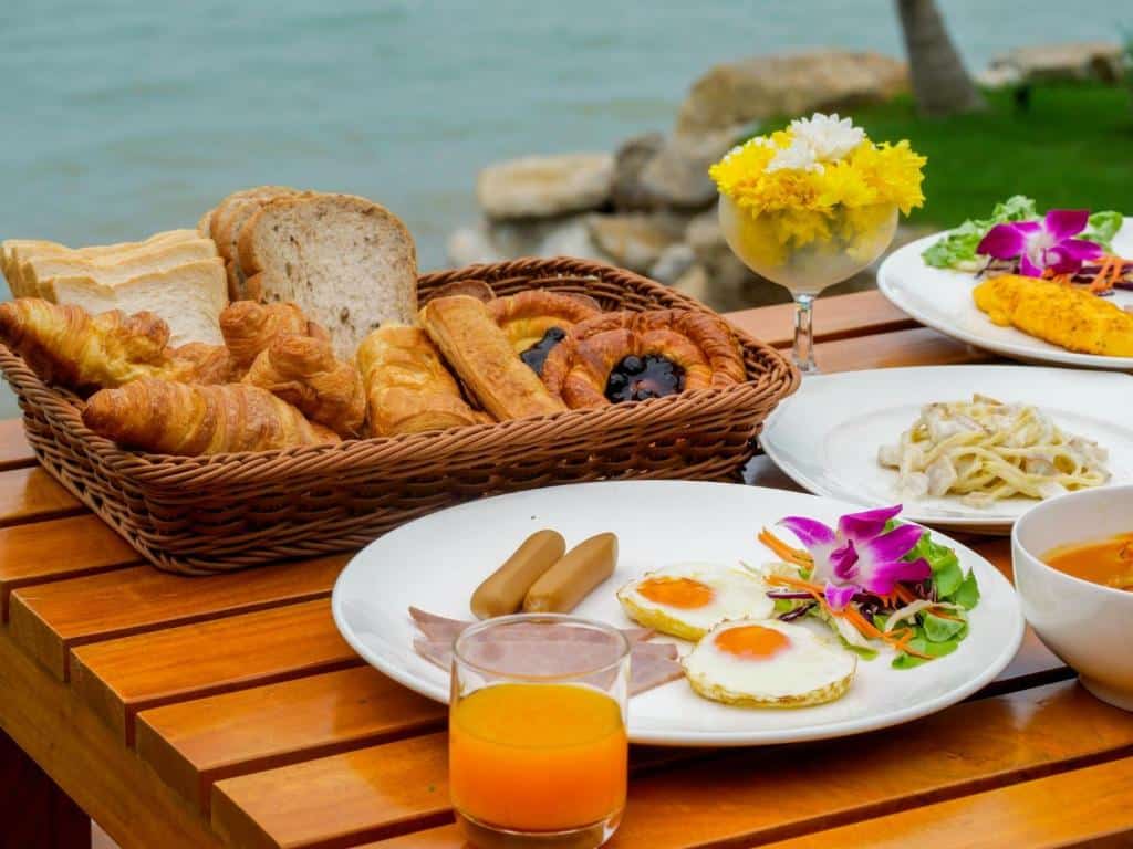 อาหารเช้ากลางแจ้งบนโต๊ะไม้ริมทะเล ประกอบด้วยตะกร้าขนมปังนานาชนิด จานพร้อมไข่ ไส้กรอก สลัด และน้ำส้มหนึ่งแก้ว ที่พักขนอมแบบครอบครัวติดทะเล