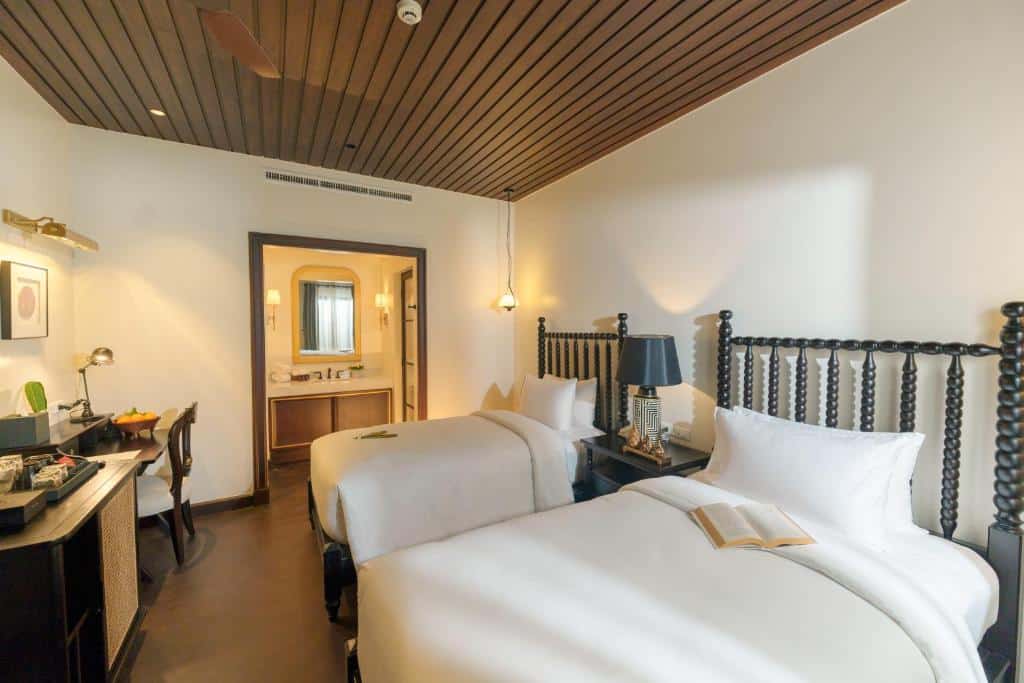 ห้องพักหรูในโรงแรมราชบุรีพร้อมเตียงคู่ ผ้าปูที่นอนสีขาว เพดานไม้ โคมไฟตั้งพื้น และหนังสือที่เปิดอยู่บนเตียง ที่เที่ยวหลวงพระบาง 