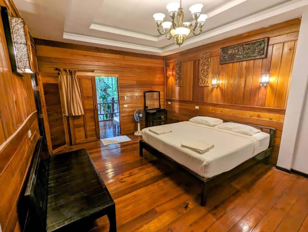 ห้องนอนแสนสบายที่บลูสกายรีสอร์ทเกาะพยามมีผนังและพื้นไม้ มีเตียงคู่ ม้านั่ง พัดลม และประตูที่นำไปสู่สวนอันเขียวชอุ่ม เกาะพยามที่พัก