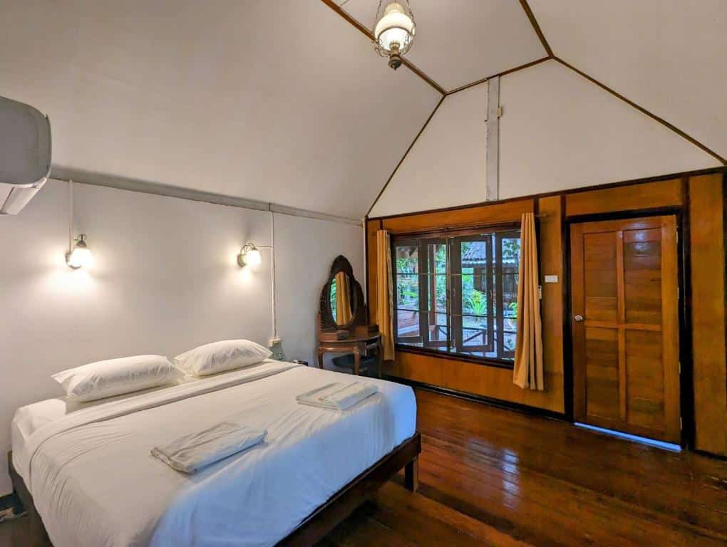 ห้องนอนแสนสบายที่มีเตียงคู่พร้อมผ้าปูที่นอนสีขาว พื้นไม้ และเฟอร์นิเจอร์ หน้าต่างบานใหญ่พร้อมผ้าม่าน และเพดานโค้งที่ Blue Sky Resort Ko Phayam เกาะพยามที่พัก