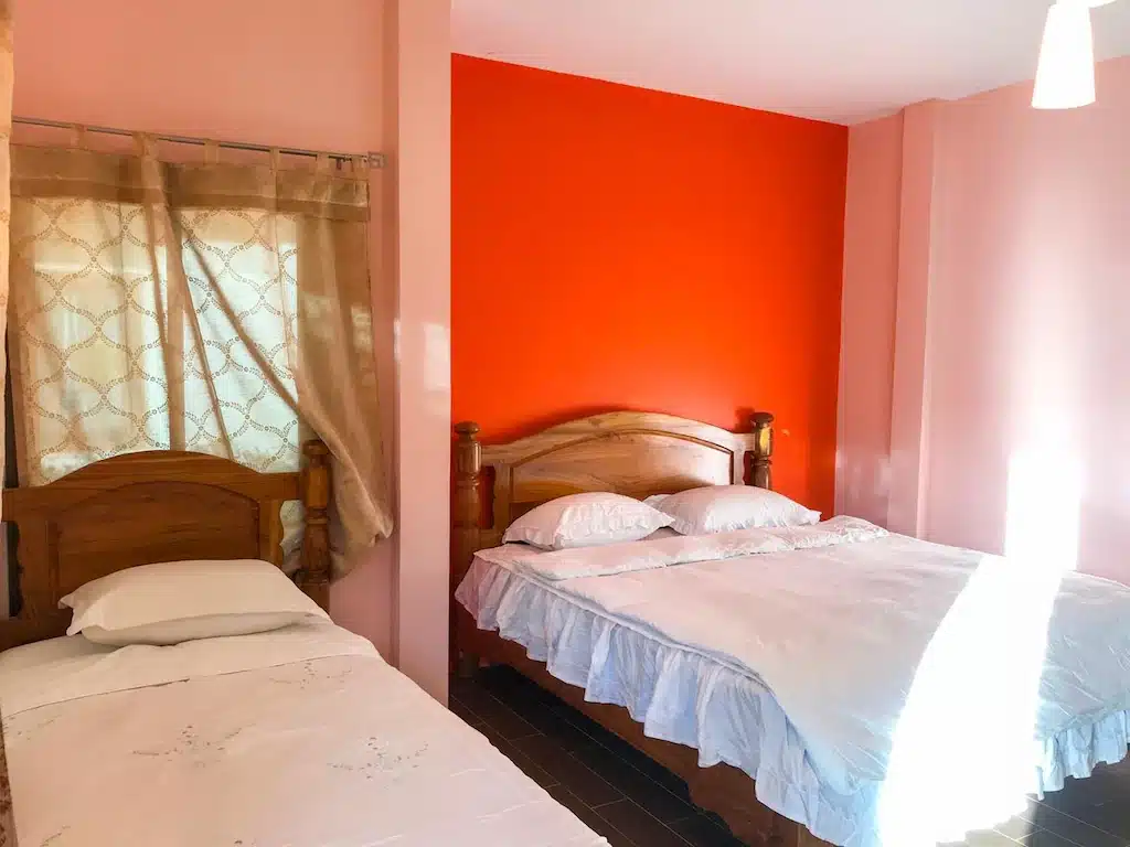 ที่พักหาดเตยงาม ห้องนอนเรียบง่ายพร้อมเตียงเดี่ยว 2 เตียงตัดกับผนังสีส้มสดใส ชุดเครื่องนอนสีขาว และหน้าต่างพร้อมผ้าม่านโปร่งอยู่ที่หาดเตยงาม