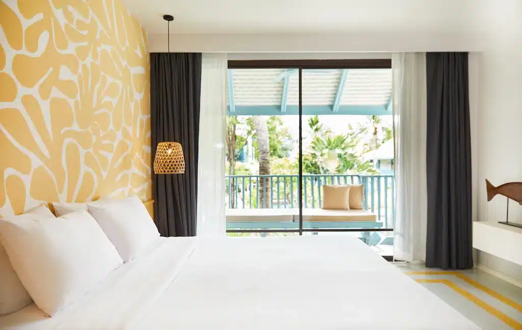 ห้องพักในโรงแรมทันสมัยที่มีเตียงขนาดใหญ่พร้อมผ้าปูที่นอนสีขาว ผนังลายสีเหลือง และประตูกระจกบานเลื่อนที่นำไปสู่ระเบียงพร้อมวิวต้นไม้ใน โรงแรมหัวหิน