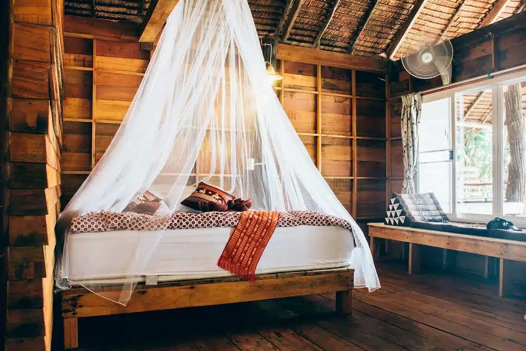ห้องนอนเคบินไม้แสนสบายที่เดอะบลูสกายรีสอร์ท เกาะพยาม มีมุ้งสีขาวพาดอยู่บนเตียง หมอนตกแต่ง และวิวภายนอกผ่านหน้าต่างบานใหญ่ เกาะพยามที่พัก