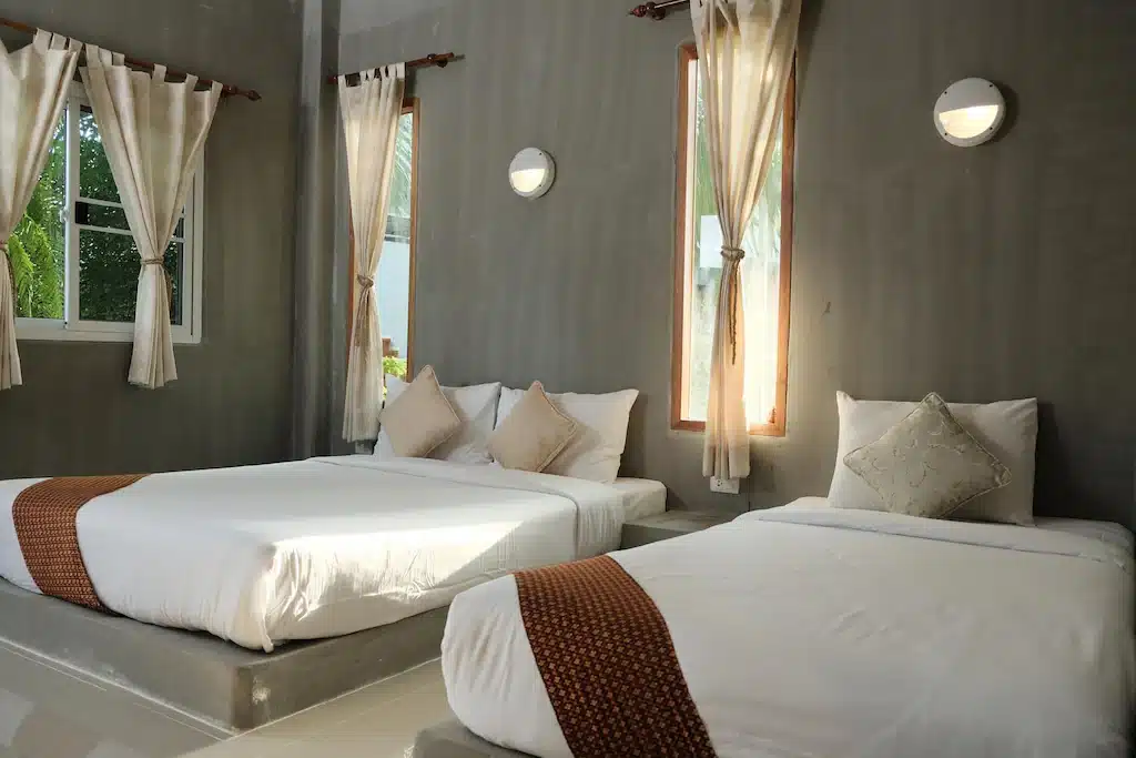 ห้องนอนทันสมัยสว่างสดใสพร้อมเตียง 2 เตียง ชุดเครื่องนอนสีขาว ผ้าคลุมสีน้ำตาล และผ้าม่านดึงบางส่วนเพื่อให้แสงแดดส่องจากทิวทัศน์อันงดงามของเมืองน่าน ที่พักขนอม