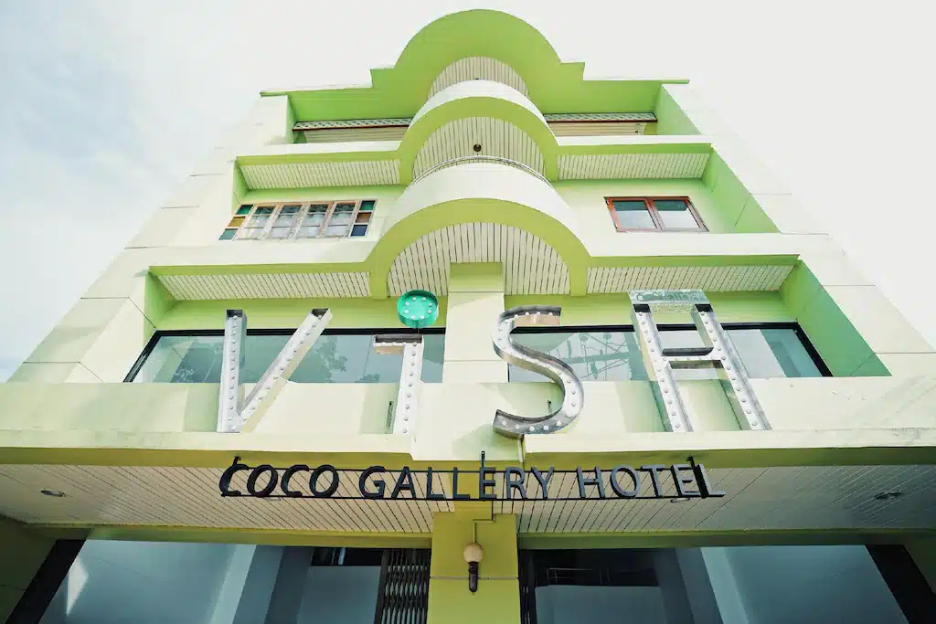 มุมมองมุมต่ำด้านหน้าโรงแรม Coco Gallery Hotel ด้วยองค์ประกอบทางสถาปัตยกรรมอาร์ตเดโคสีเขียวที่ โรงแรมขนอม โดดเด่นภายใต้ท้องฟ้าแจ่มใสในน่านที่เที่ยว
