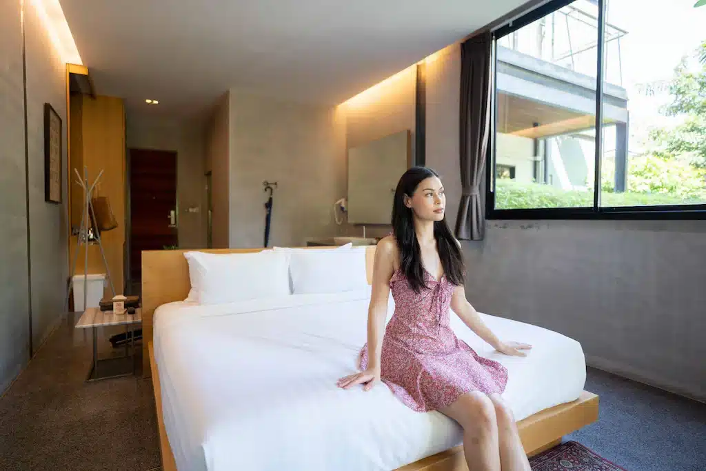 ผู้หญิงในชุดสีชมพูนั่งอยู่ขอบเตียงในห้องนอนทันสมัยที่มีหน้าต่างบานใหญ่และทิวทัศน์อันเขียวขจีด้านนอก เที่ยวเกาะเสม็ด
