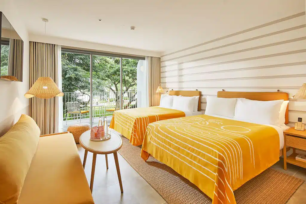 ห้องพัก โรงแรมหัวหิน ทันสมัยสว่างสดใส มีเตียง 2 เตียงพร้อมผ้าคลุมเตียงสีส้ม พื้นที่นั่งเล่นขนาดเล็ก หน้าต่างบานใหญ่พร้อมวิวสวน และผนังลายทาง รีสอร์ทหัวหิน