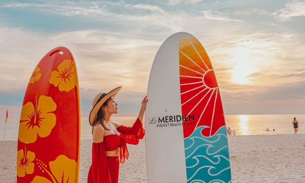 ผู้หญิงในชุดสีแดงและหมวกกำลังพิงกระดานโต้คลื่นที่ชายหาดในจังหวัดพังงาช่วงพระอาทิตย์ตก โดยมีกระดานสีสันสดใสอีกกระดานอยู่ข้างๆ พูลวิลล่าพังงา